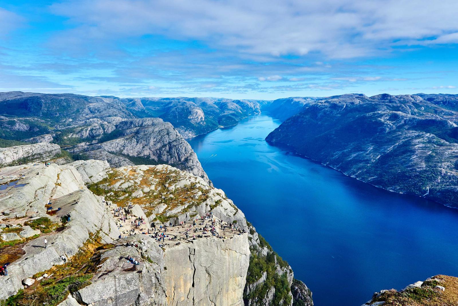 Aerial photograph of Preikestolen cliff over the Lysefjord in Norway, with a bright sky, deep blue water, and tiny people standing on the rock